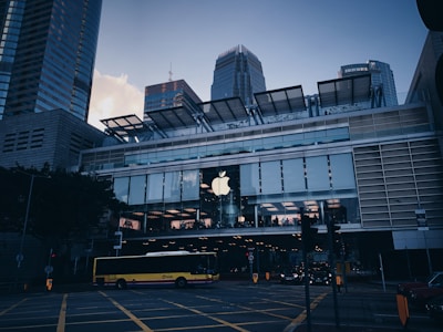 A modern cityscape featuring a prominent commercial building with a large Apple logo on the facade. The structure is composed of glass and metal, with a spacious second-floor window showcasing the interior lighting. A yellow bus is seen crossing the street, surrounded by traffic lights and road markings. Skyscrapers tower in the background, adding to the urban atmosphere.