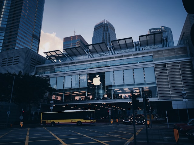 A modern cityscape featuring a prominent commercial building with a large Apple logo on the facade. The structure is composed of glass and metal, with a spacious second-floor window showcasing the interior lighting. A yellow bus is seen crossing the street, surrounded by traffic lights and road markings. Skyscrapers tower in the background, adding to the urban atmosphere.