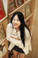 A person with long black hair and a smiling expression holds a book while standing next to a bookshelf. The shelf contains various colorful books, primarily cookbooks, arranged neatly. The individual wears a white shirt and a plaid skirt, and the setting appears to be cozy and inviting.