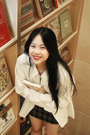 A person with long black hair and a smiling expression holds a book while standing next to a bookshelf. The shelf contains various colorful books, primarily cookbooks, arranged neatly. The individual wears a white shirt and a plaid skirt, and the setting appears to be cozy and inviting.