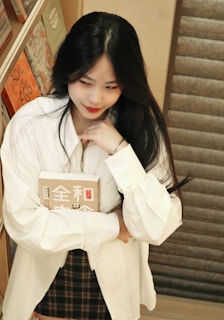 A woman with long dark hair is standing near a wooden bookshelf. She is wearing a white shirt and a plaid skirt, holding a book with Asian characters on the cover. Her expression is thoughtful as she gazes downward.