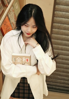 A woman with long dark hair is standing near a wooden bookshelf. She is wearing a white shirt and a plaid skirt, holding a book with Asian characters on the cover. Her expression is thoughtful as she gazes downward.