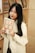 A woman with long black hair is holding a book in front of a bookshelf. She is wearing a white coat and smiling subtly. The bookshelf behind her contains various books with different colored spines.