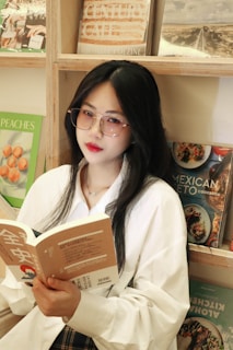 A woman with long black hair and glasses is reading a book. She is wearing a white shirt and is seated in front of a bookshelf filled with various books, including titles related to cuisine.
