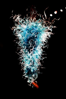 A stunning glass sculpture with intricate swirling patterns and vivid blue and white hues. The artwork exudes a sense of fluidity and complexity, with delicate tendrils extending outward. It is displayed against a dark background, highlighting its luminous and ethereal quality.