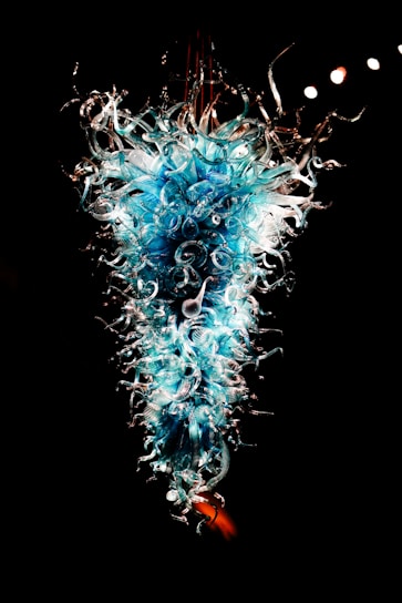 A stunning glass sculpture with intricate swirling patterns and vivid blue and white hues. The artwork exudes a sense of fluidity and complexity, with delicate tendrils extending outward. It is displayed against a dark background, highlighting its luminous and ethereal quality.