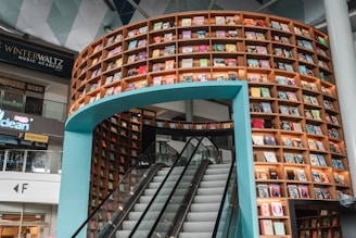 A curved bookshelf filled with colorful books creates an artistic backdrop for an escalator in a modern indoor space. The structure is part of a commercial area, as indicated by adjacent signs for a music academy and retail stores. Warm lighting enhances the visual appeal of the books and the surrounding architecture.