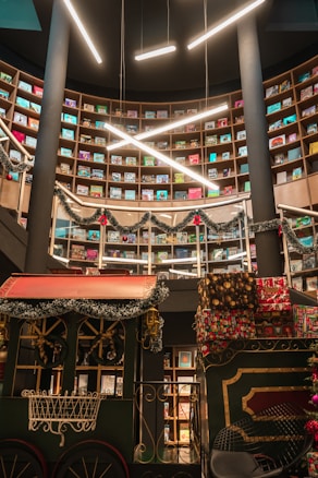 A library or bookstore with multiple levels of rounded bookshelves filled with brightly colored books. The upper area is decorated with festive garlands and red ornaments. In the foreground, there is a decorative train car adorned with garlands and presents, suggesting a holiday theme. The area is well-lit with modern linear lights hanging from the ceiling.