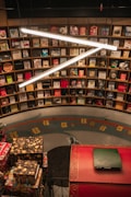 A bookstore filled with a variety of books on wooden shelves arranged in a circular pattern. Bright, white overhead lights create a modern ambiance. Decorative elements such as wrapped gifts are placed on a red, ornately designed ledge.