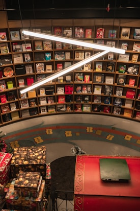A bookstore filled with a variety of books on wooden shelves arranged in a circular pattern. Bright, white overhead lights create a modern ambiance. Decorative elements such as wrapped gifts are placed on a red, ornately designed ledge.