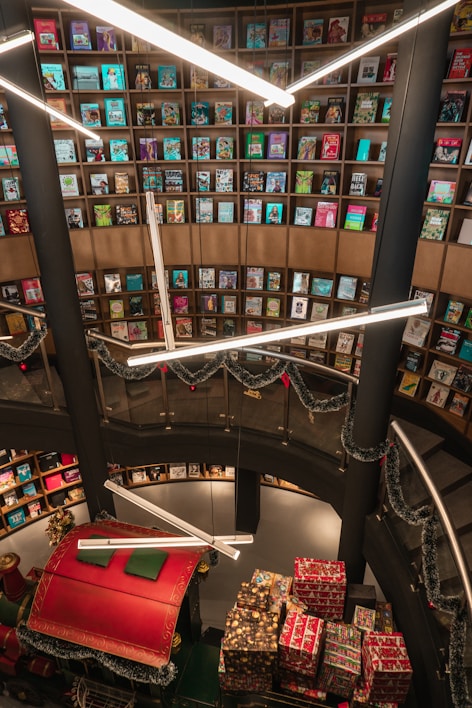 A library or bookstore with a tall, circular bookshelf filled with colorful books stretching from the floor to the ceiling. Bright, modern lighting fixtures hang overhead, casting light on the surroundings. Holiday decorations, including garlands and a section of a sleigh filled with wrapped presents, add a festive touch to the space.