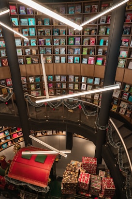 A library or bookstore with a tall, circular bookshelf filled with colorful books stretching from the floor to the ceiling. Bright, modern lighting fixtures hang overhead, casting light on the surroundings. Holiday decorations, including garlands and a section of a sleigh filled with wrapped presents, add a festive touch to the space.