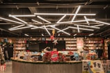 A well-lit bookstore interior with shelves full of books and a variety of products on display. A counter is visible at the center with two people working behind it, surrounded by gift-wrapped items and decorations. Modern geometric lighting fixtures hang from the ceiling, adding a contemporary feel to the space. There are signs and promotional materials scattered around, adding to the festive atmosphere.