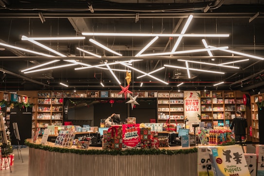A well-lit bookstore interior with shelves full of books and a variety of products on display. A counter is visible at the center with two people working behind it, surrounded by gift-wrapped items and decorations. Modern geometric lighting fixtures hang from the ceiling, adding a contemporary feel to the space. There are signs and promotional materials scattered around, adding to the festive atmosphere.