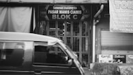 A black and white image depicting the front of a market stall in a local bazaar with a sign reading 'Pasar Manis Ciamis Blok C'. A vehicle is passing by in the foreground. Signs displaying various menu items are visible on the right side.