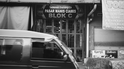 A black and white image depicting the front of a market stall in a local bazaar with a sign reading 'Pasar Manis Ciamis Blok C'. A vehicle is passing by in the foreground. Signs displaying various menu items are visible on the right side.