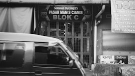 A black and white image depicting the front of a market stall in a local bazaar with a sign reading 'Pasar Manis Ciamis Blok C'. A vehicle is passing by in the foreground. Signs displaying various menu items are visible on the right side.