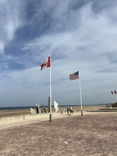 A scenic view of a coastline with flags of Canada and the United States prominently displayed on flagpoles. In the background, a beach extends to the horizon under a partly cloudy sky. There are people walking near a modern monument near the shore with artistic, metallic structures. Other flags are visible in the distance.