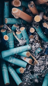 A collection of cigarette butts and ashes, predominantly featuring broken, turquoise-colored cigarette filters among gray ash. The image has a gritty, used look, conveying a sense of pollution and waste.