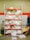 A storage shelving unit filled with large white plastic bags, possibly containing materials or waste. The shelves are painted red and the background consists of a brick wall with a red stripe. Various cleaning equipment, such as brooms and dustpans, are placed on the left side. On the right, there are cardboard sheets leaning against the wall.