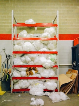 A storage shelving unit filled with large white plastic bags, possibly containing materials or waste. The shelves are painted red and the background consists of a brick wall with a red stripe. Various cleaning equipment, such as brooms and dustpans, are placed on the left side. On the right, there are cardboard sheets leaning against the wall.