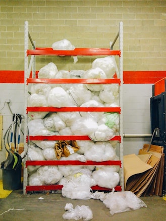 A storage shelving unit filled with large white plastic bags, possibly containing materials or waste. The shelves are painted red and the background consists of a brick wall with a red stripe. Various cleaning equipment, such as brooms and dustpans, are placed on the left side. On the right, there are cardboard sheets leaning against the wall.