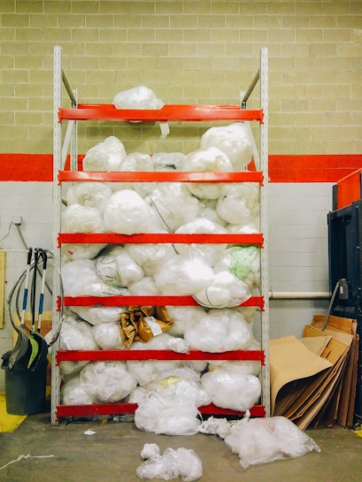 A storage shelving unit filled with large white plastic bags, possibly containing materials or waste. The shelves are painted red and the background consists of a brick wall with a red stripe. Various cleaning equipment, such as brooms and dustpans, are placed on the left side. On the right, there are cardboard sheets leaning against the wall.