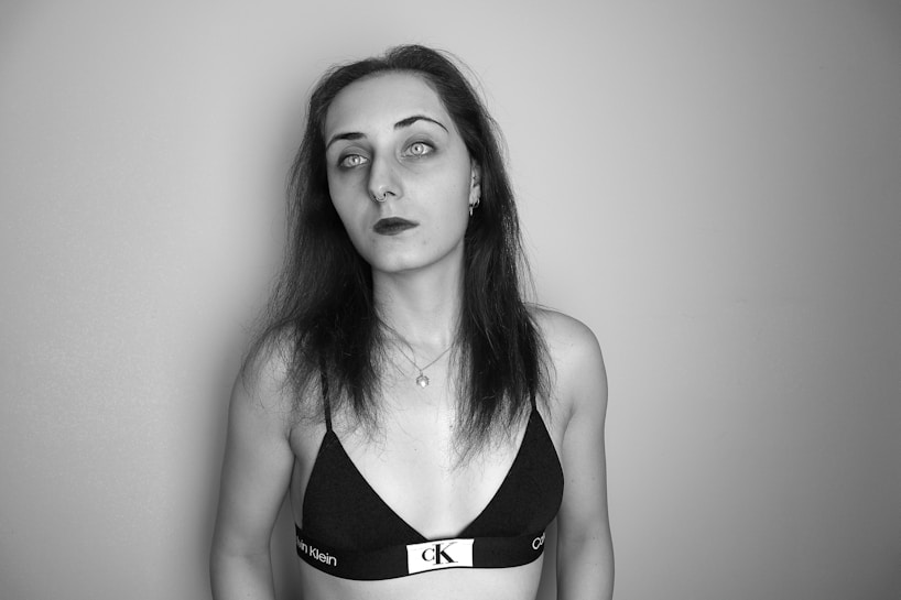 A monochrome portrait of a person with long dark hair, wearing a branded black top and a necklace. The person has a neutral expression and is positioned against a plain background.