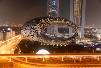 A futuristic building with a unique elliptical shape is illuminated against a city skyline at night. The structure is covered with stylized Arabic calligraphy and stands prominently against the backdrop of the urban landscape, which includes other high-rise buildings and busy roads lit by streetlights.