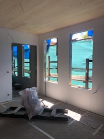 A room under construction with unfinished walls, exposed wooden beams, and large windows covered by blue tarp. A pile of construction materials, including plastic sheet and wooden boards, is situated on the floor. Sunlight filters through the windows, highlighting the interior.