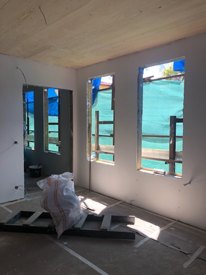 A room under construction with unfinished walls, exposed wooden beams, and large windows covered by blue tarp. A pile of construction materials, including plastic sheet and wooden boards, is situated on the floor. Sunlight filters through the windows, highlighting the interior.