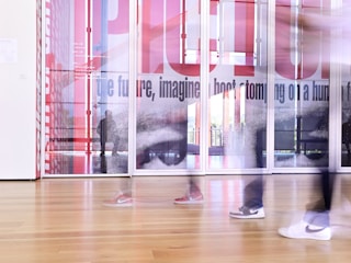 A modern interior space featuring blurred figures walking past large glass panels with artistic designs and text. The floor is wooden, and the overall scene is bright, creating a dynamic and contemporary atmosphere.