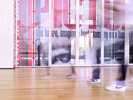 A modern interior space featuring blurred figures walking past large glass panels with artistic designs and text. The floor is wooden, and the overall scene is bright, creating a dynamic and contemporary atmosphere.