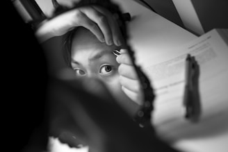 A person is seen using tweezers to pluck an eyebrow while looking into a handheld round mirror. The focus is on one eye, and the scene includes a book and a pen in the background. The image is in black and white, creating a classic and focused composition.