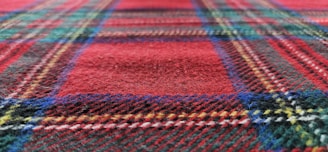 A close-up view of a textured fabric with a plaid pattern, featuring intersecting lines in various colors such as red, green, blue, and yellow. The texture appears soft and fuzzy.