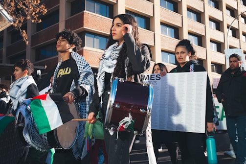 A group of people participate in an outdoor protest, holding Palestinian flags and playing drums. One person carries a poster with text. They wear keffiyehs and dress in casual clothing, while standing in front of a modern building with reflective windows.