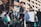 A group of people participate in an outdoor protest, holding Palestinian flags and playing drums. One person carries a poster with text. They wear keffiyehs and dress in casual clothing, while standing in front of a modern building with reflective windows.