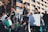 A group of people participate in an outdoor protest, holding Palestinian flags and playing drums. One person carries a poster with text. They wear keffiyehs and dress in casual clothing, while standing in front of a modern building with reflective windows.