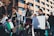 A group of people participate in an outdoor protest, holding Palestinian flags and playing drums. One person carries a poster with text. They wear keffiyehs and dress in casual clothing, while standing in front of a modern building with reflective windows.