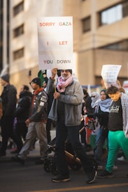 A group of people are participating in a protest or demonstration. One individual in the foreground is holding a large sign reading 'SORRY GAZA I LET YOU DOWN'. The participants are dressed in casual attire, some wearing keffiyeh scarves, and are walking along a city street. Background elements include blurred buildings and additional protest signs.