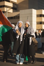 Two individuals wearing traditional scarves stand together, holding a large flag with red, green, black, and white colors. Both are smiling and wearing long coats. They appear to be part of a larger gathering or protest in an urban setting with tall buildings in the background.