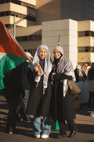 Two individuals wearing traditional scarves stand together, holding a large flag with red, green, black, and white colors. Both are smiling and wearing long coats. They appear to be part of a larger gathering or protest in an urban setting with tall buildings in the background.