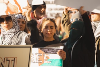 A group of individuals are gathered, holding signs and wearing clothing associated with a protest or demonstration setting. Some wear patterned headscarves and sunglasses, while one person in the forefront is holding a sign and has eyes closed, appearing focused or contemplative.