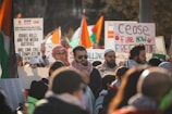 A group of people gathered for a protest, holding signs with various messages related to political issues. Some individuals are wearing headscarves and sunglasses. There are numerous flags in the background, suggesting a focus on a specific region or cause.