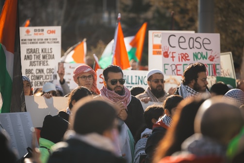 A group of people gathered for a protest, holding signs with various messages related to political issues. Some individuals are wearing headscarves and sunglasses. There are numerous flags in the background, suggesting a focus on a specific region or cause.