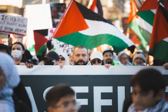 A group of people are gathered for a demonstration, holding up banners and flags with red, white, green, and black colors, indicative of a Palestinian theme. Among the crowd, one man is prominently visible, appearing determined and focused.