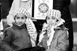 Two children wearing traditional black and white checkered scarves and jackets stand closely together. They are in front of an adult holding a sign that has an image and the words 'Free Palestine.' The background includes parts of other people, suggesting a public gathering or protest.