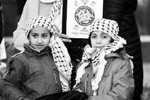 Two children wearing traditional black and white checkered scarves and jackets stand closely together. They are in front of an adult holding a sign that has an image and the words 'Free Palestine.' The background includes parts of other people, suggesting a public gathering or protest.