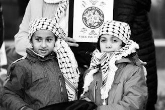 Two children wearing traditional black and white checkered scarves and jackets stand closely together. They are in front of an adult holding a sign that has an image and the words 'Free Palestine.' The background includes parts of other people, suggesting a public gathering or protest.