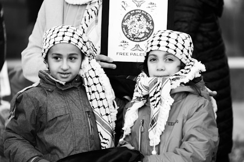 Two children wearing traditional black and white checkered scarves and jackets stand closely together. They are in front of an adult holding a sign that has an image and the words 'Free Palestine.' The background includes parts of other people, suggesting a public gathering or protest.